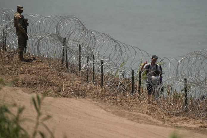 Migrantes caminan entre alambre de púas frente a un agente tras haber cruzado el río Bravo. 