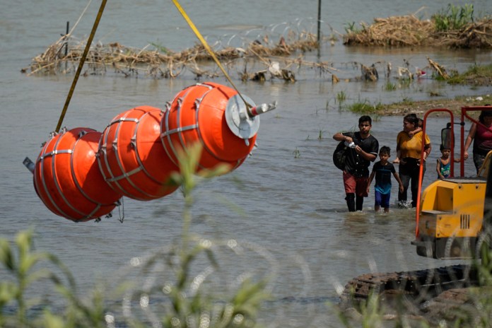 Migrantes observan unas boyas grandes que serán utilizadas como valla fronteriza en el río Bravo, a la altura de Eagle Pass, 