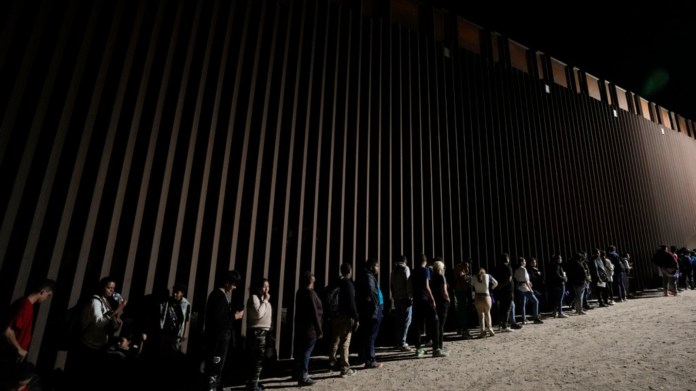 Varias personas se forman junto a un muro fronterizo mientras esperan solicitar asilo tras cruzar la frontera desde México