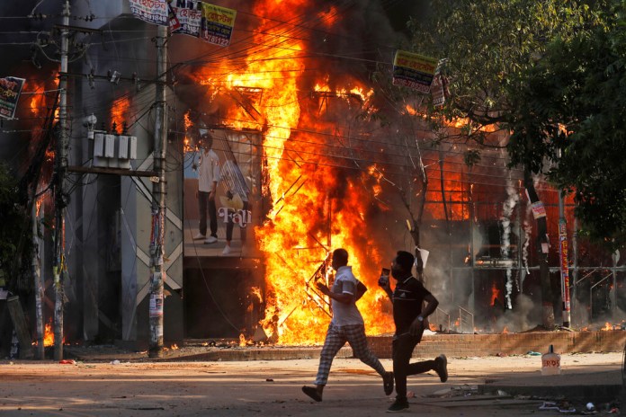 Unos hombres corren frente a un centro comercial que fue incendiado durante una manifestación contra la primera ministra Sheikh Hasina y su gobierno, en Dacca, Bangladesh, el domingo 4 de agosto de 2024. (Foto AP/Rajib Dhar) Photo Details