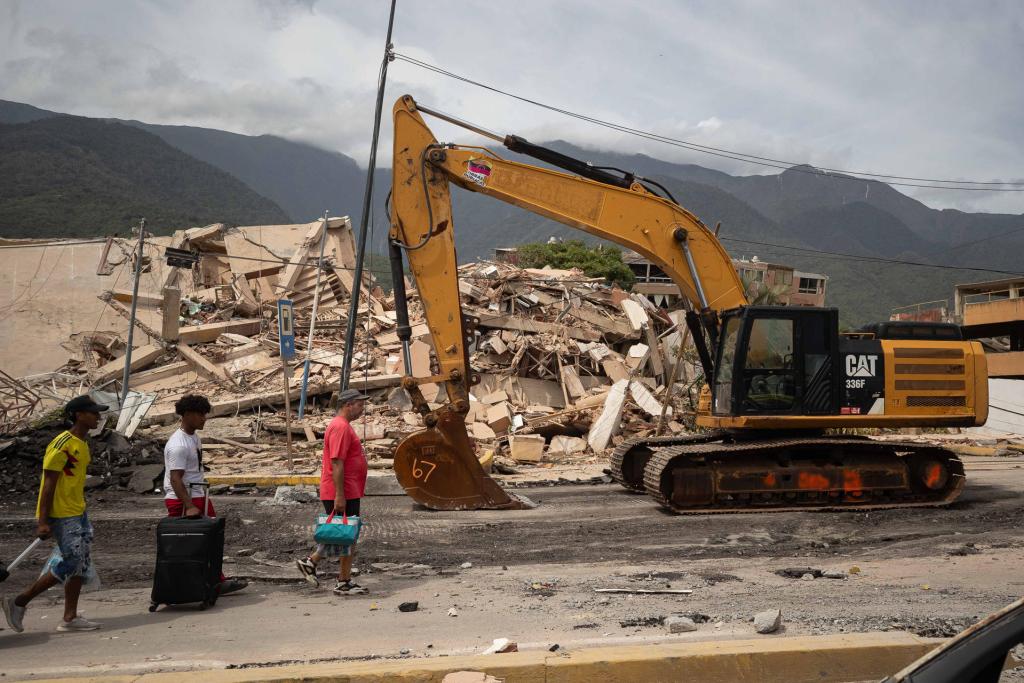 Personas caminan frente a una máquina que realiza labores de remoción de escombros este jueves, en La Guaira. Foto La Hora: EFE